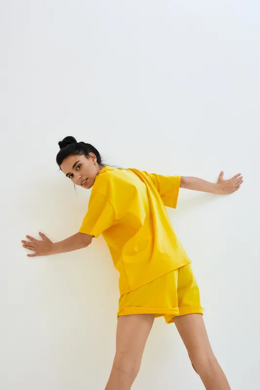 Young woman in yellow sporty outfit leans against white wall, showing dynamic pose and casual style with natural lighting in minimalist indoor setting.