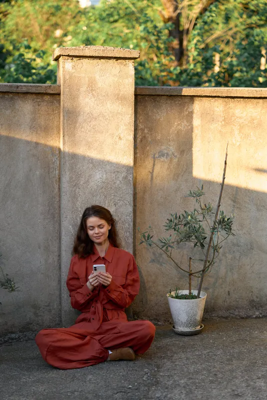 A woman in a red dress sits cross legged beside a stone wall, looking at her phone in a sunlit courtyard. A relaxed lifestyle moment, calm focus, and quiet contemplation