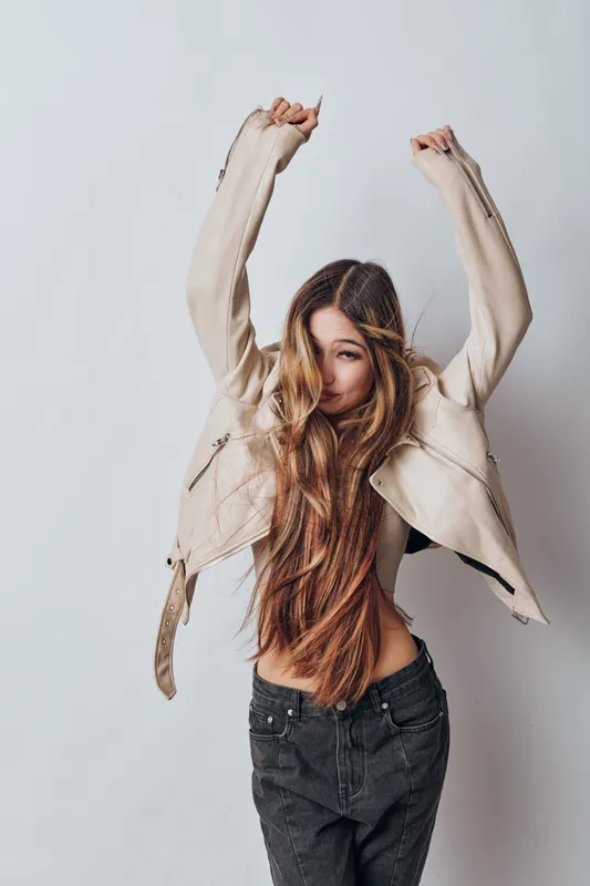 Woman with long hair stretching arms wearing beige jacket and black jeans against plain background, casual fashion and relaxed pose for lifestyle portrait photography.