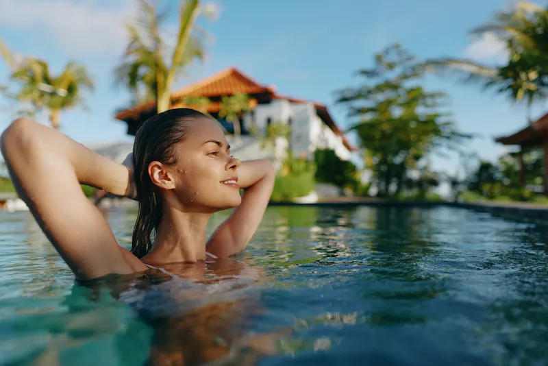 A young woman relaxes in a sunlit pool with arms behind her head, eyes closed, smiling softly, while palm trees and a resort building glow in the background, conveying serenity and wellness.