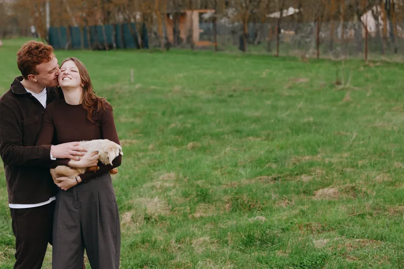 Young couple embraces outdoors while holding a small goat in their arms, sharing a tender moment on a grassy field with natural spring colors and relaxed casual clothing.