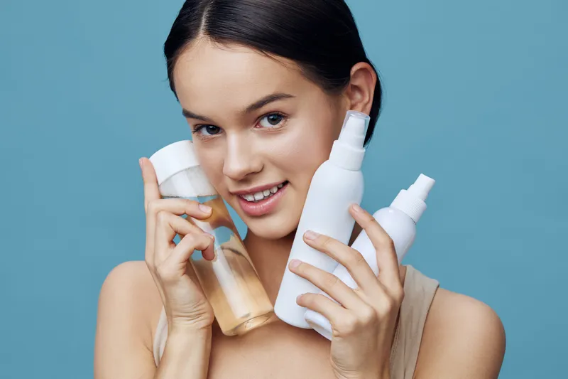 A young woman holding skincare products with a bright smile, showcasing healthy skin, in a minimalistic blue background, emphasizing beauty and self care