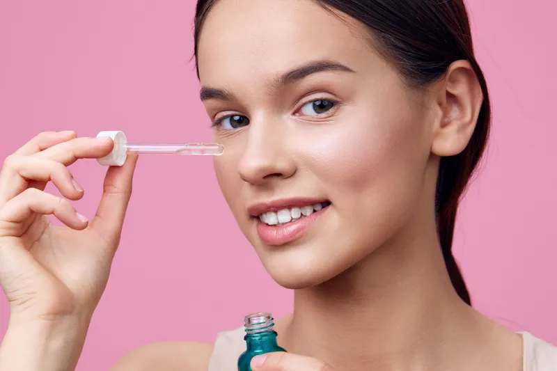 Young woman applying serum to her face, smiling confidently against a pink background, highlighting beauty and skincare concepts in a fresh and vibrant setting
