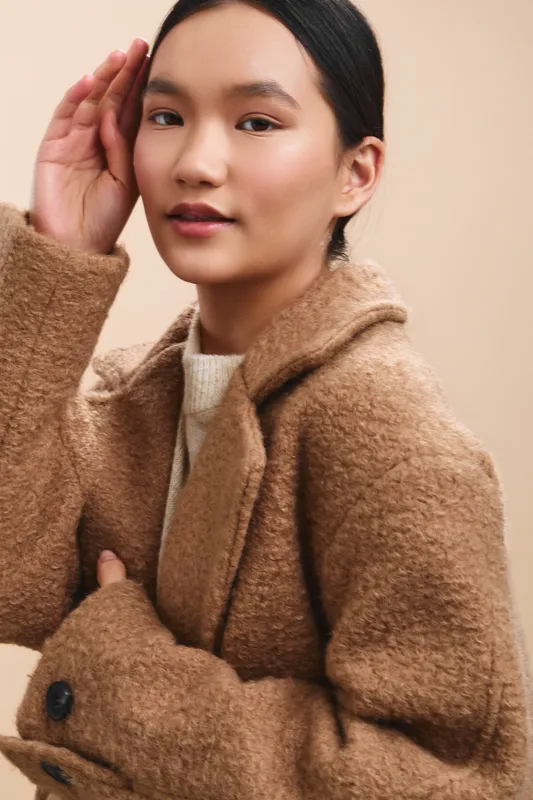 Asian woman in a cozy tan coat stands in a soft studio setting, conveying elegance and warmth through poised posture and natural expression, inviting attention and suggesting approachable