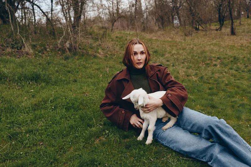 A woman in casual clothes sits on a grassy field with a young goat kid, capturing a calm outdoor lifestyle moment, gentle bonding, and connection with nature.