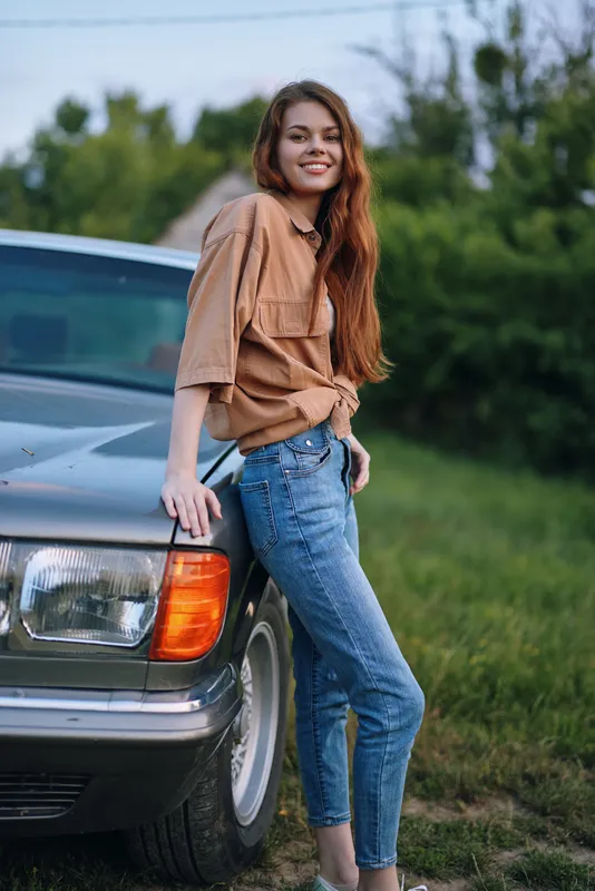 Young woman in casual attire smiling by an old car in a green outdoor setting perfect for summer. Stylish and relaxed vibe captured in a vibrant scene.