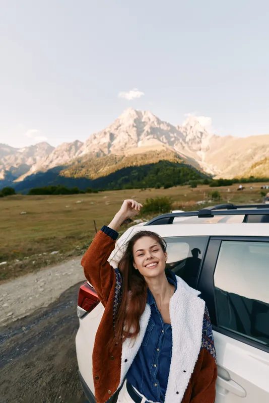 Woman leans from car with a bright smile beside a mountain road during travel, enjoying the landscape and nature. Casual jacket, relaxed portrait capturing outdoor journey and scenic freedom.