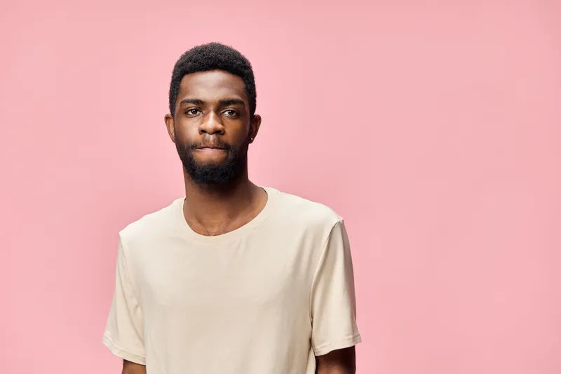 Young African man in a neutral t shirt with a contemplative expression against a pink backdrop, conveying introspection and thoughtfulness, ideal for lifestyle themes