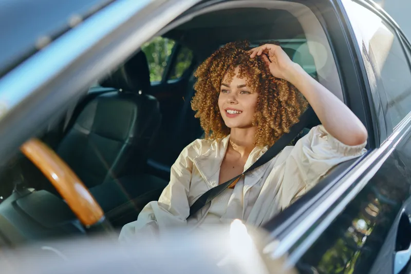 Smiling woman with curly hair sitting in black car wearing light jacket and seatbelt, enjoying sunny day with relaxed and happy expression.