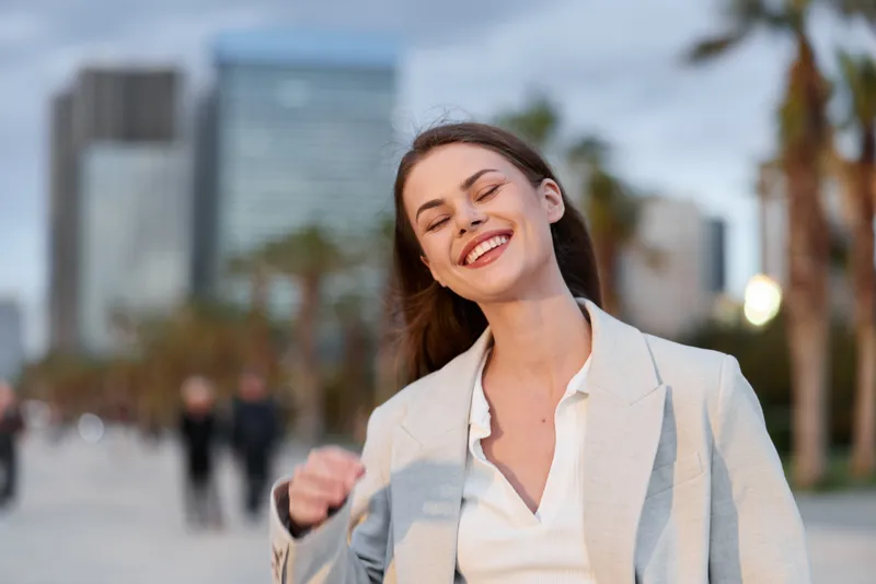 Happy young woman in beige blazer smiling outdoors in urban business district with cityscape background and soft natural lighting