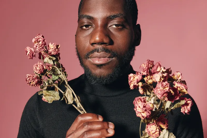 African man with beard holding dried pink flowers in both hands against pink background in creative conceptual visual style portrait with serious expression and black sweater.