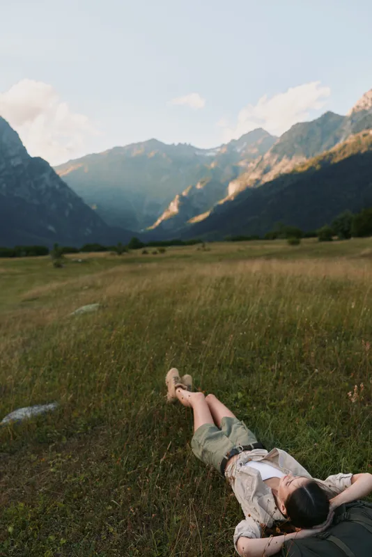 Woman lifestyle concept with young woman lying on grass field surrounded by mountains under clear sky enjoying peaceful nature and relaxation in outdoor environment.