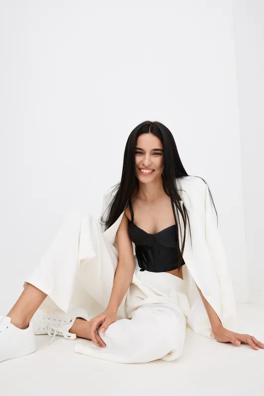 Smiling woman with long black hair wears white suit and black top, sitting relaxed on white floor in bright minimalist studio, showcasing casual modern fashion.
