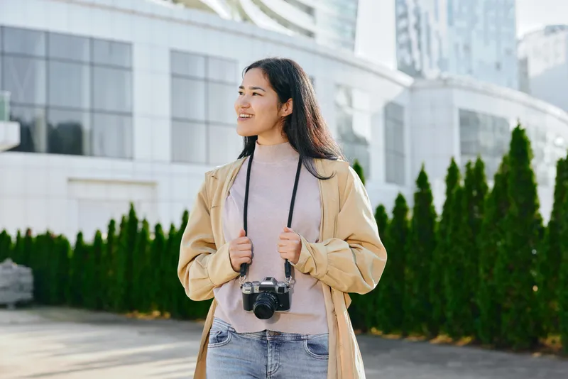 Woman with camera smiling outdoors in an urban setting, casual fashion, bright daylight, street photography vibe, stylish jacket, modern architecture, candid moment.