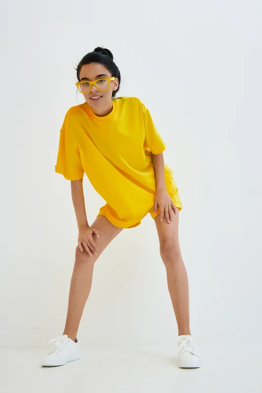 Young woman in yellow oversized t-shirt and yellow glasses posing on white background, expressing casual style and joyful attitude with sporty white sneakers.