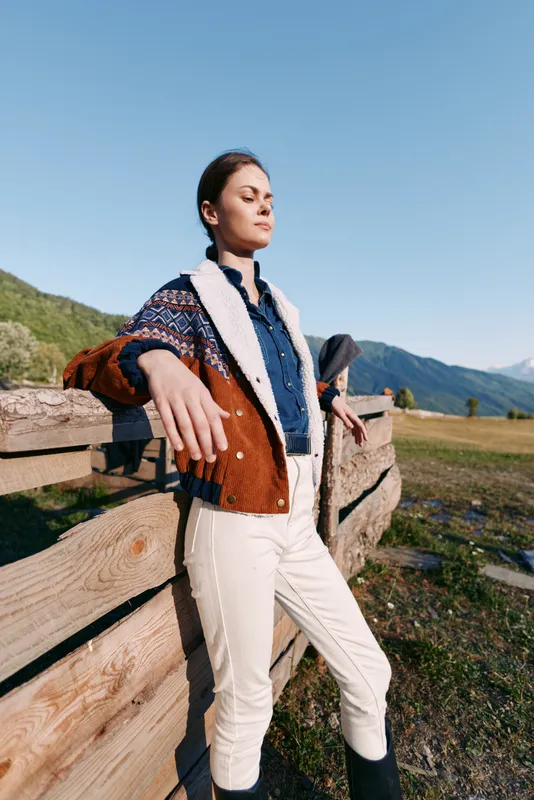 Woman leaning on wooden fence in mountains countryside wearing patterned cardigan and denim shirt, white trousers and boots, eyes closed enjoying warm sunlight and fresh air.
