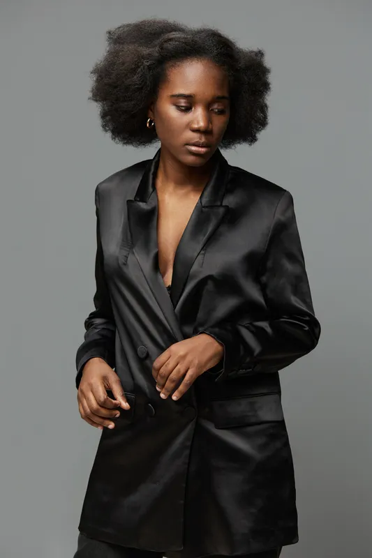 Elegant black female model in satin blazer, showcasing confidence and grace against a neutral gray background, perfect for fashion and beauty concepts