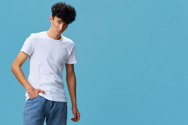 Young man wearing a white t-shirt and blue jeans standing against a plain blue background with a casual and relaxed pose, showcasing casual fashion style.