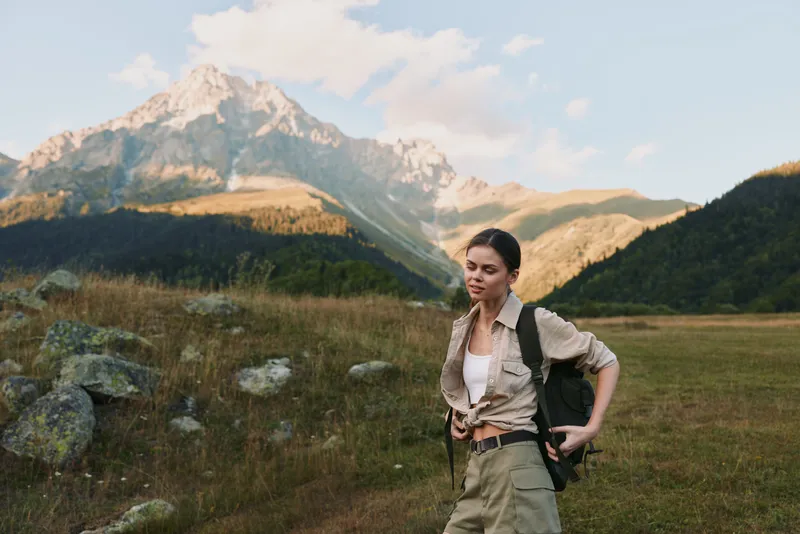 A brunette woman with a backpack stands on a rocky mountain trail, pausing to breathe as the expansive valley and craggy peaks glow under bright daylight, conveying calm and a sense of adventure.