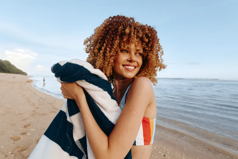 Cheerful woman on a sunny beach, wearing a striped swimsuit, hugging a towel, smiling toward the camera, breeze in hair, relaxed pose, bright natural colors, calm sea background
