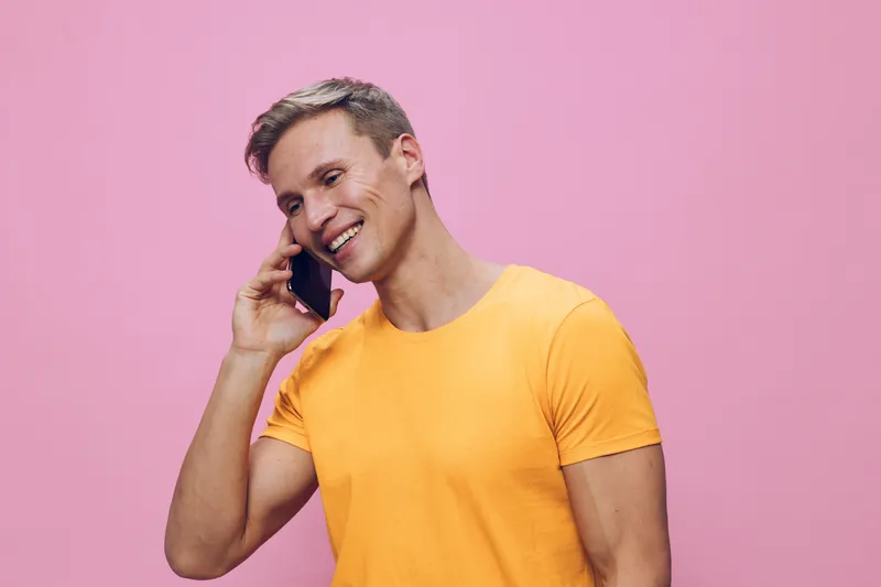 Smiling man talking on mobile phone against pink background, wearing yellow t-shirt, casual, cheerful expression, modern lifestyle, studio portrait, bright colors, youthful vibe, happiness, communicat