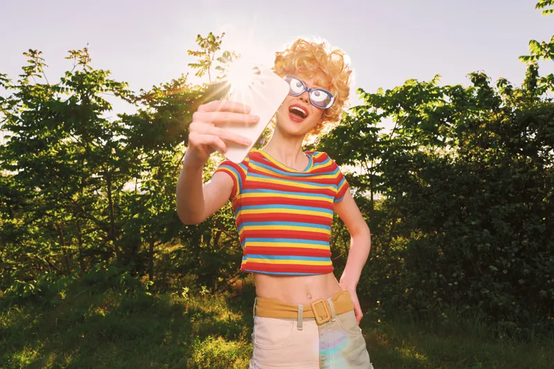 Woman lifestyle shot with retro film color features a joyful woman taking a selfie outdoors in bright sun, striped top, sunglasses, carefree vibe, golden hour glow.