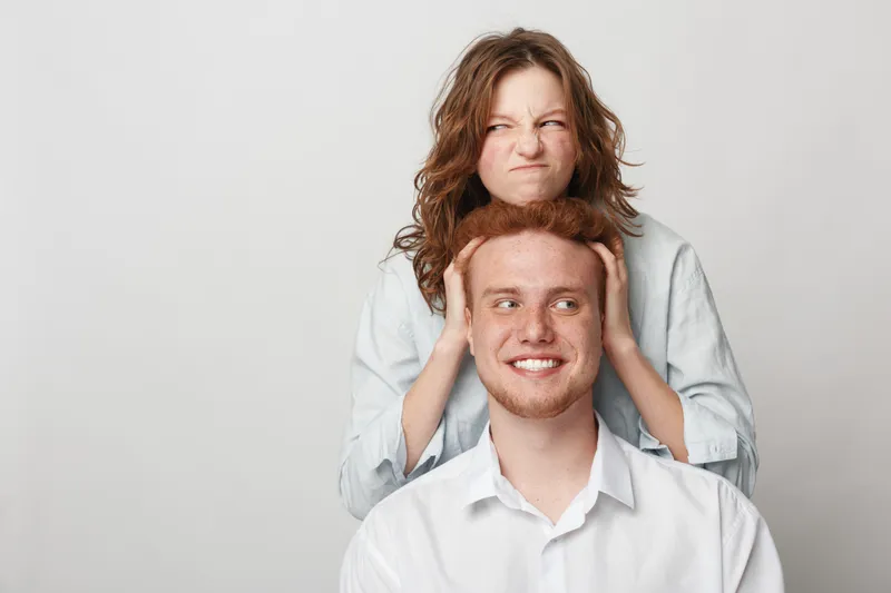 Redhead woman playfully pulling redhead man’s hair while he smiles. Both wear casual white shirts against plain background. Concept of fun, friendship, and playful interaction between young adults.