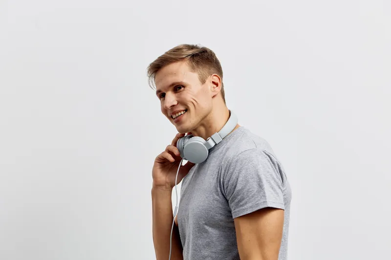 Smiling man with short brown hair wearing casual gray t-shirt holding headphones, looking cheerful, friendly, and relaxed, studio portrait on plain white background, lifestyle concept