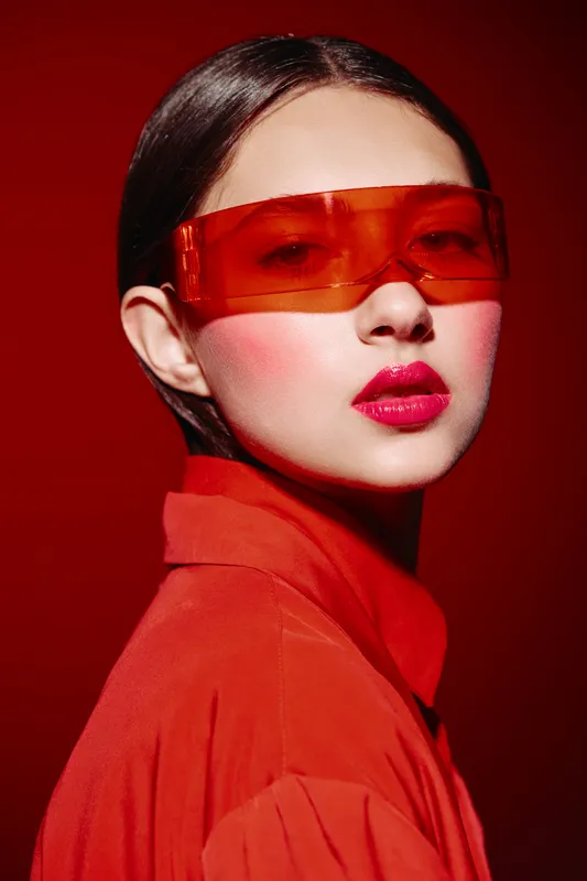 Fashion model in bright red outfit and translucent red visor poses against red background. Makeup includes bold pink lipstick and strong blush. Concept of beauty and fashion creativity.