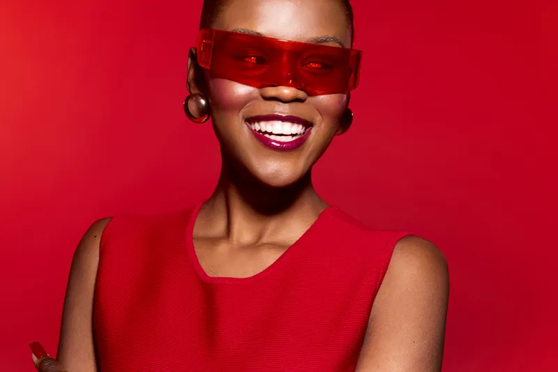 Smiling African American woman with dark skin wearing red sleeveless dress and red sunglasses on red background. Concept of fashion, style, confidence, and vibrant look.