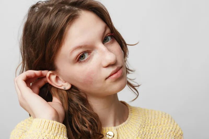 Young woman with natural skin and light makeup looks at camera with soft expression. She wears a yellow sweater and poses against a plain white background. Concept of beauty and simplicity.