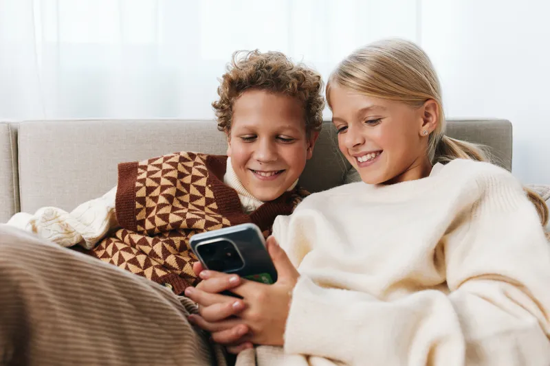Authentic moment of playful learning and family lifestyle as two children share a cozy screen moment on the sofa, smiling together in a warm, relaxed setting.