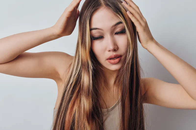 Woman with long hair and natural makeup posing with hands on head, showing confident expression in studio with plain background for beauty and fashion concept.