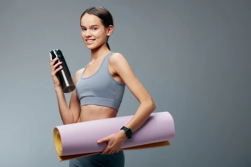 Woman fitness model in athletic wear holds a water bottle and a rolled yoga mat, showcasing an active lifestyle, workout routine and gym readiness.