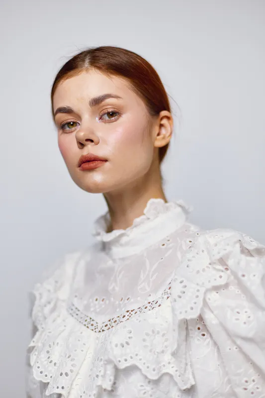 Elegant young woman with a thoughtful expression, wearing a white ruffled blouse, against a soft, neutral background, conveying a sense of beauty and serenity