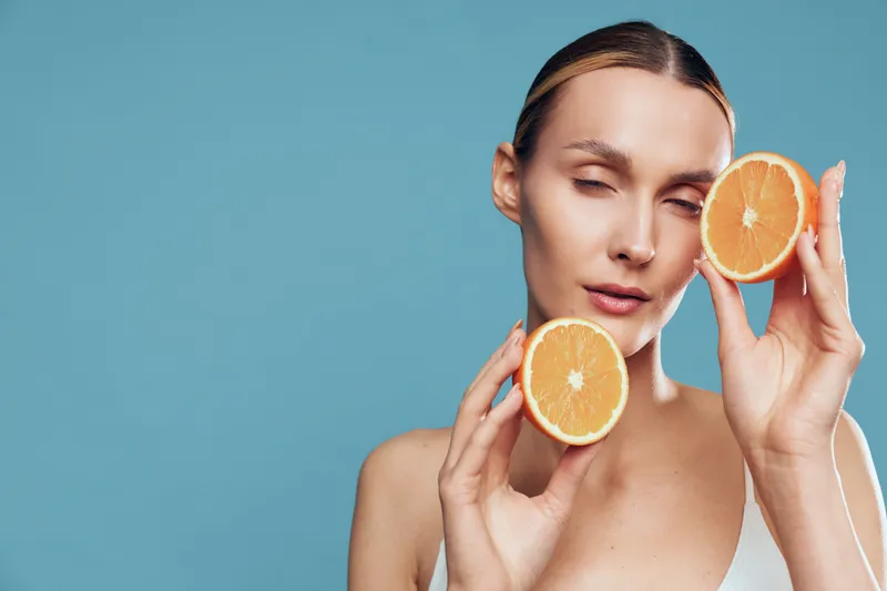 Young woman with clear skin holds two halves of a fresh orange near her face against a blue background expressing freshness and natural beauty with closed eyes.