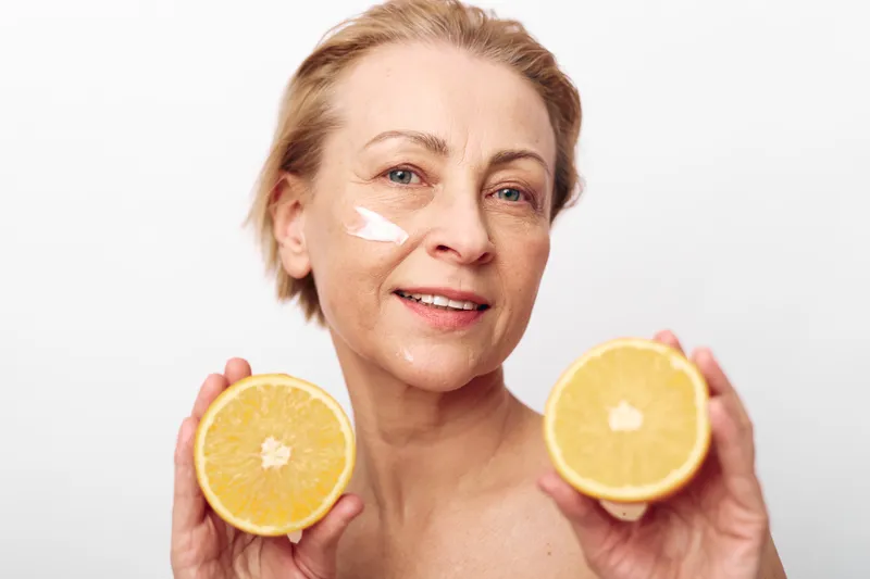 Mature woman with short hair applies white cream on cheek holding two halves of fresh orange. Healthy skincare concept with citrus fruits and natural beauty on white background.