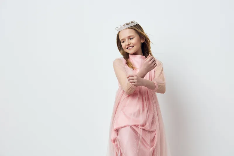 Happy girl in a flowing pink dress with a crown, showcasing playful emotions in a bright and clean environment, perfect for children s fashion and celebrations