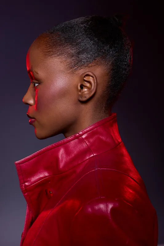 Profile of a black woman with dark skin wearing a red leather jacket. Studio portrait with dramatic lighting on a dark background. Concept for fashion, style, and beauty.
