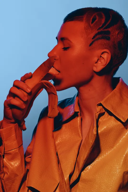 Young woman with artistic shaved hairstyle holding and smelling a banana, wearing a stylish tan jacket against a blue background with warm red lighting, modern creative portrait.