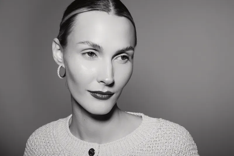 Portrait of a young Caucasian woman with fair skin wearing hoop earrings and a knitted sweater, looking away thoughtfully on a neutral background.
