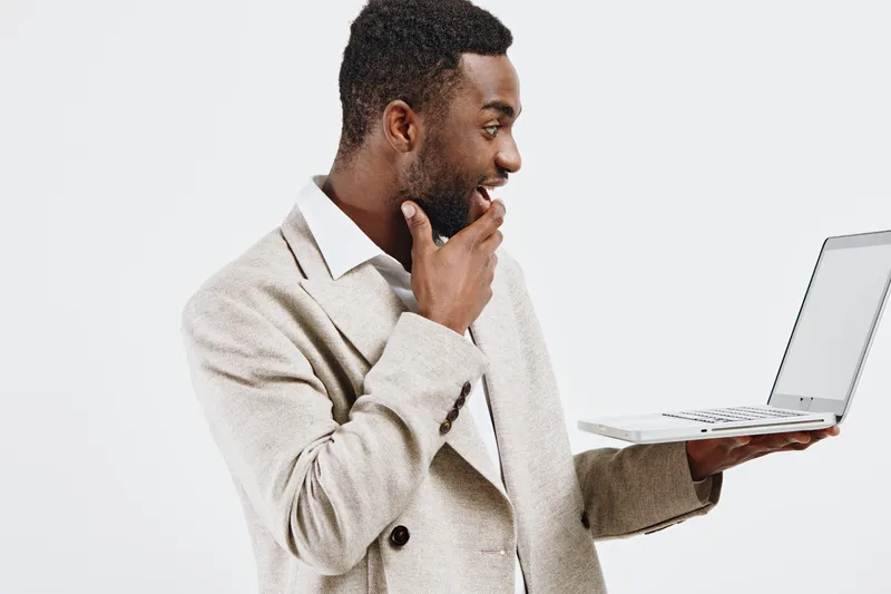 Happy young man with dark skin holding a laptop and looking at it thoughtfully, wearing a beige blazer, standing against a plain white background, studio portrait. People lifestyle concept, modern tec