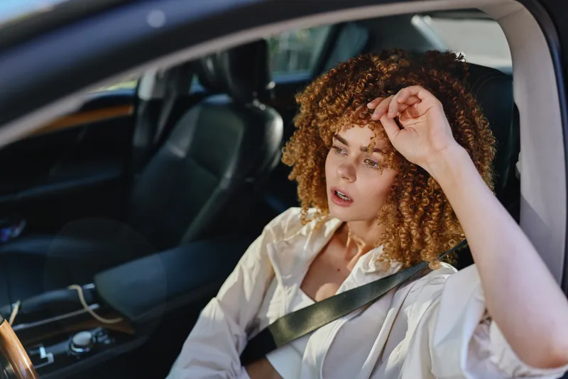 young woman with curly hair sitting in car wearing seatbelt, looking tired and wiping forehead, casual white shirt, sunlight through window