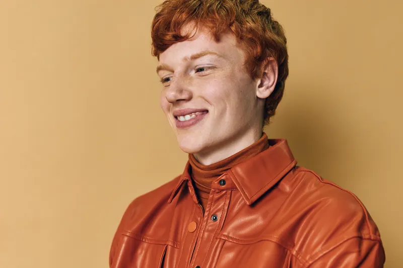 Young man with red hair wearing orange leather jacket smiles and looks down with happy expression on solid beige background in closeup portrait.