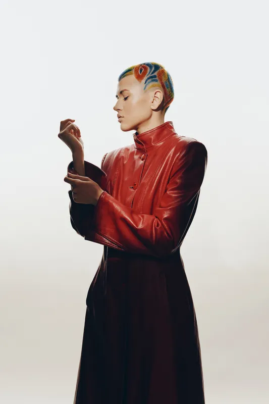 Woman with colorful short hair wearing a red leather jacket, standing in profile against a light background with a contemplative expression.