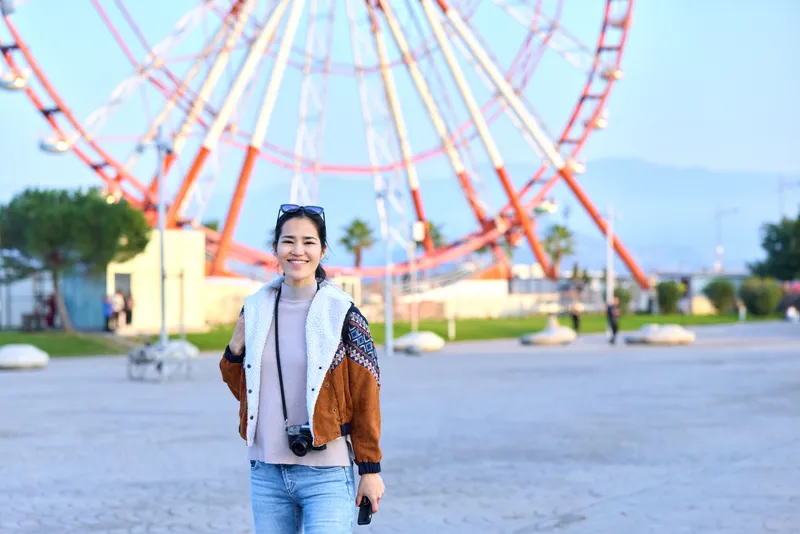 Woman wearing casual jacket and jeans stands in an outdoor amusement park beside a towering ferris wheel, capturing a cheerful travel mood with vivid daylight colors
