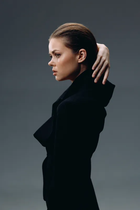 Profile of a serious woman in a black coat with sleek hair, posing against a dark background with focused expression.