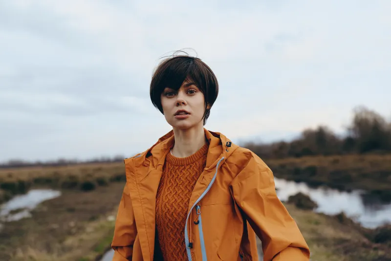 Woman lifestyle portrait outdoors in orange jacket and brown sweater, standing on a windy countryside path under cloudy sky, calm thoughtful expression, natural light, travel fashion