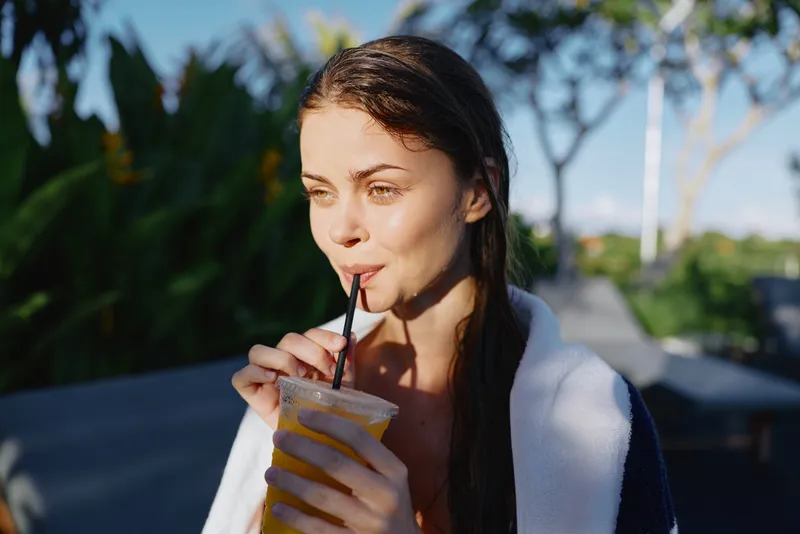 Relaxed woman sipping a smoothie outdoors beside a pool, towel draped over shoulders. Bright natural light conveys leisure, lifestyle, and a refreshing self care moment.