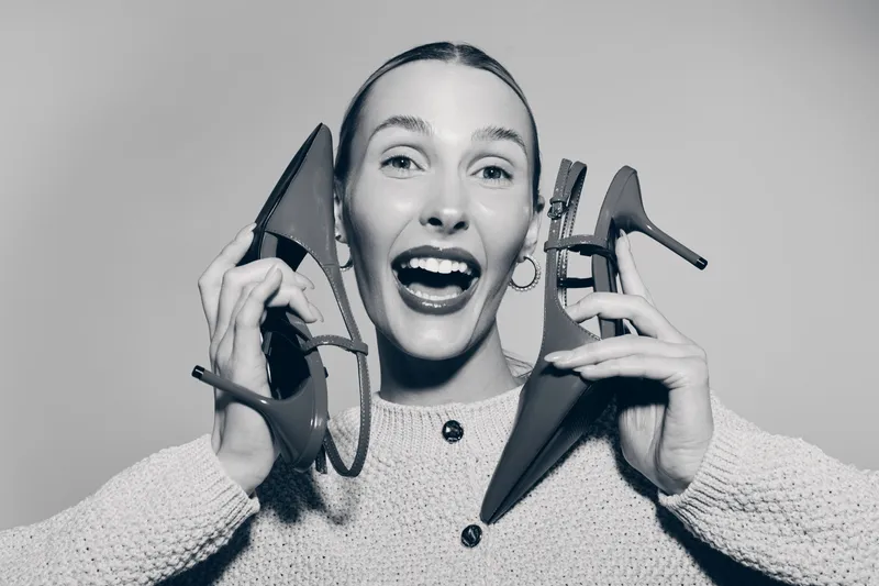 Happy caucasian woman with light skin holds high heel shoes near face, smiling widely with excitement in studio.